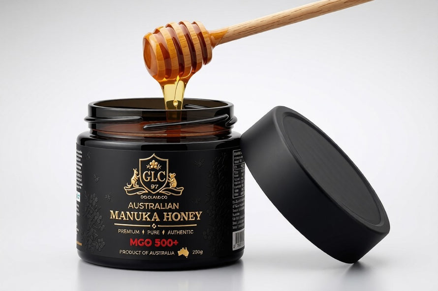 Thick amber Australian Manuka honey lifting from an open Goldland Co. MGO 500+ jar on a wooden dipper, with the black crest-embossed lid resting alongside on a light grey backdrop.